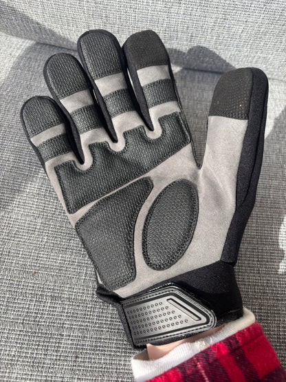 A single work glove shown flat against a gray textured fabric background. The glove is primarily light gray with black textured panels on the fingers, palm, and wrist strap, plus a small red-and-white plaid cuff visible at the bottom edge. Safety Tuff gloves with finger guards (finger caps).