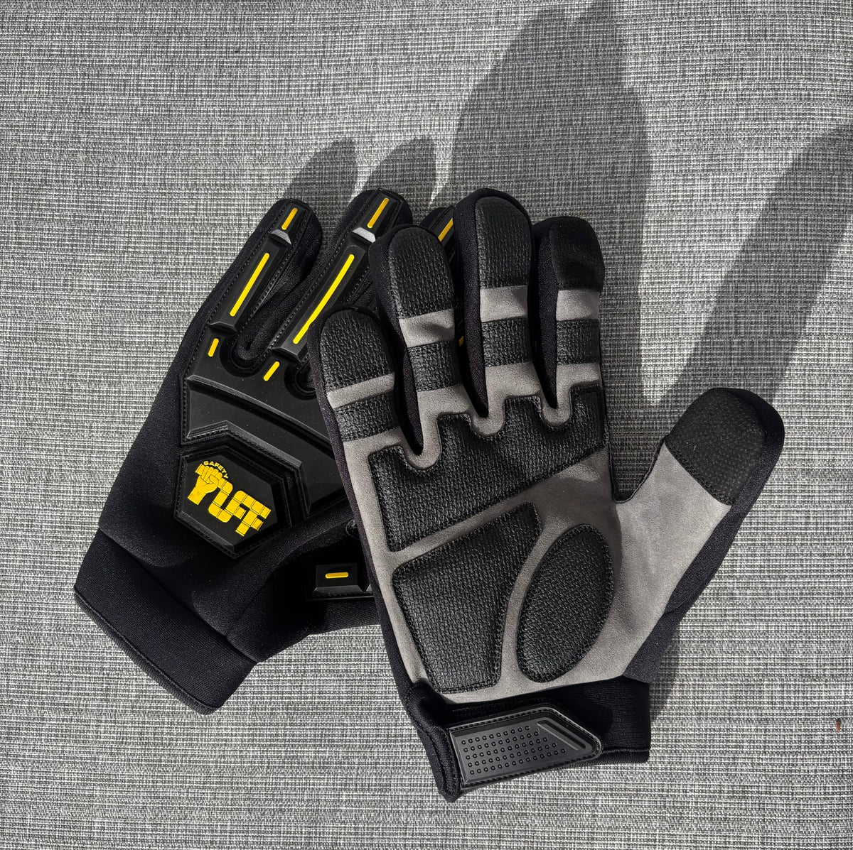 A pair of black and gray protective gloves with yellow accents, including a visible 'TUFF' logo on the back, laid flat on a gray textured surface.Safety Tuff gloves with finger guards (finger caps)